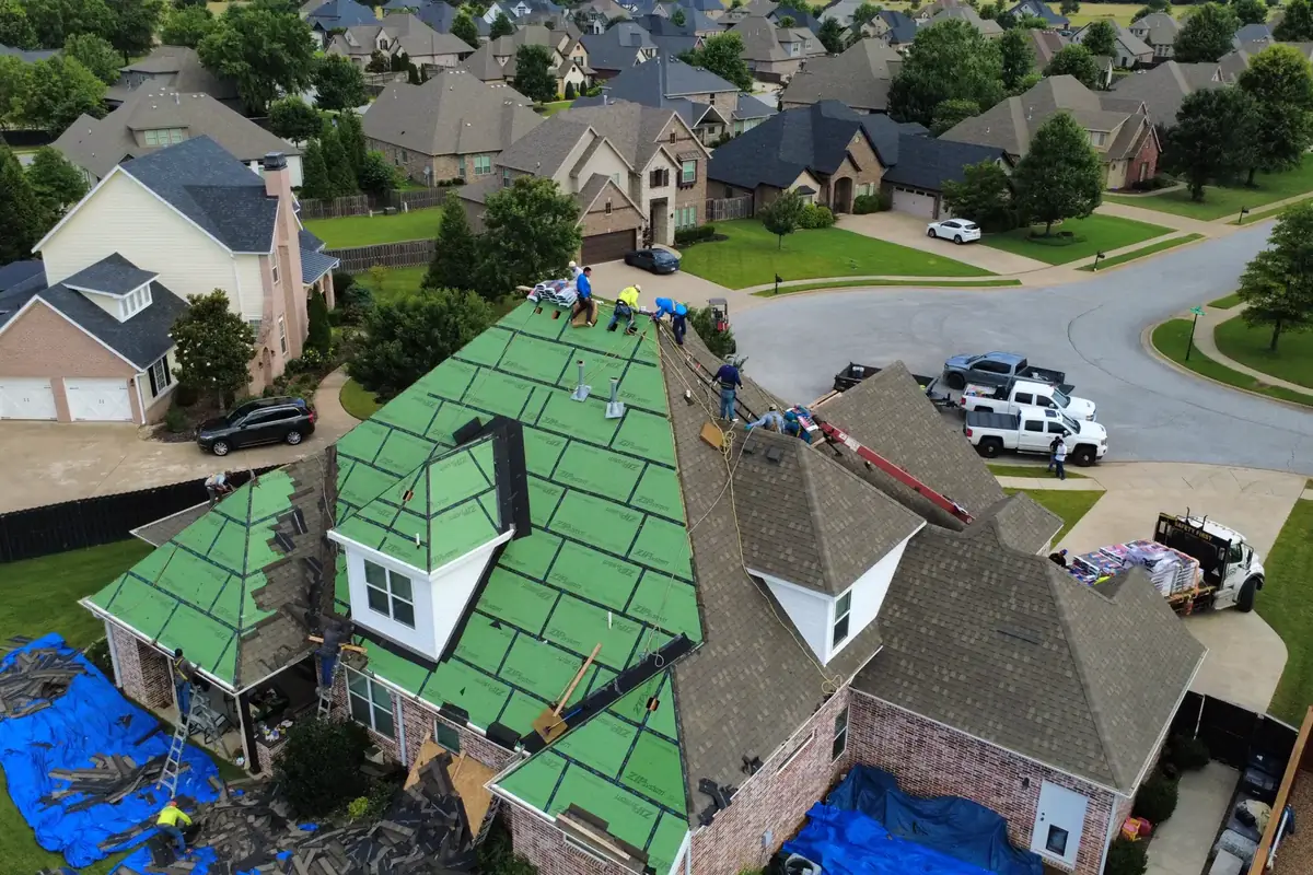 Aerial view of a roof replacement in progress: old shingles coming off, the green roof deck showing, and tarps catching debris in the yard